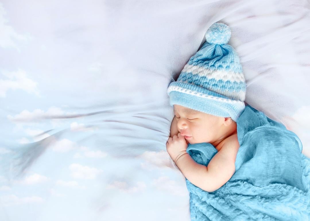 A sleeping baby bundled in a blue beanie with a pom pom and a blue blanket..